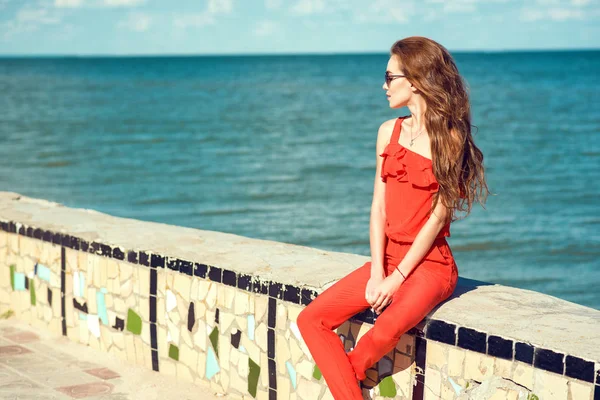 Joven hermosa mujer elegante glam con el pelo ondulado largo con mono rojo coral y gafas de sol oscuras de moda sentado en el viejo parapeto de mosaico de hormigón en la playa y mirando a un lado —  Fotos de Stock