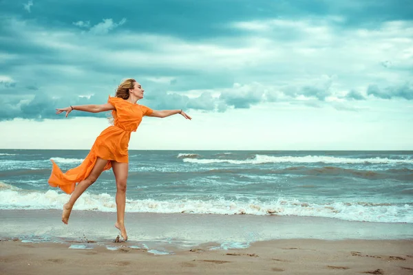 在橙色迷你礼服用飞行火车跳舞赤脚在暴风雨的海面的湿沙上美丽苗条金发女子肖像 — 图库照片
