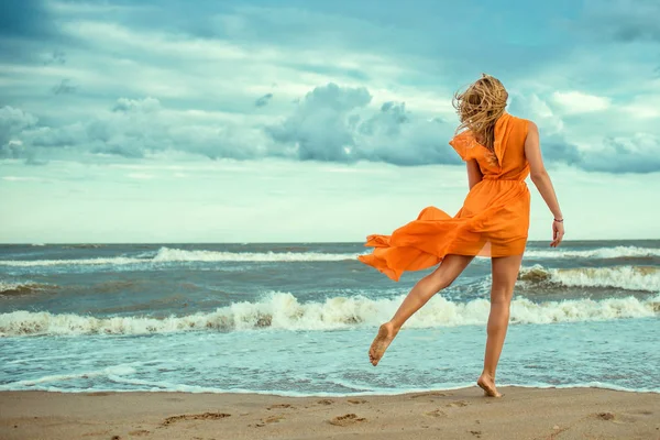 Retrato de una hermosa mujer rubia delgada en mini vestido naranja con tren volador bailando descalzo sobre la arena mojada en el mar tormentoso —  Fotos de Stock