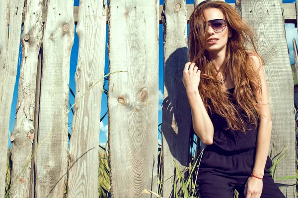 年轻漂亮的模特，身穿黑色连身裤及时尚太阳眼镜站在旧的破旧栅栏木板制成的长长的栗色头发 — 图库照片