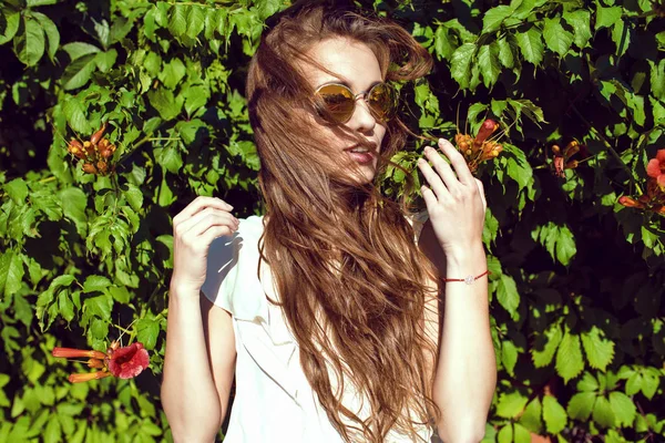 Mujer hermosa joven con el pelo largo castaño soplado por el viento usando gafas de sol de espejo redondas de moda de pie en el seto de enredadera Virginia y mirando a un lado —  Fotos de Stock