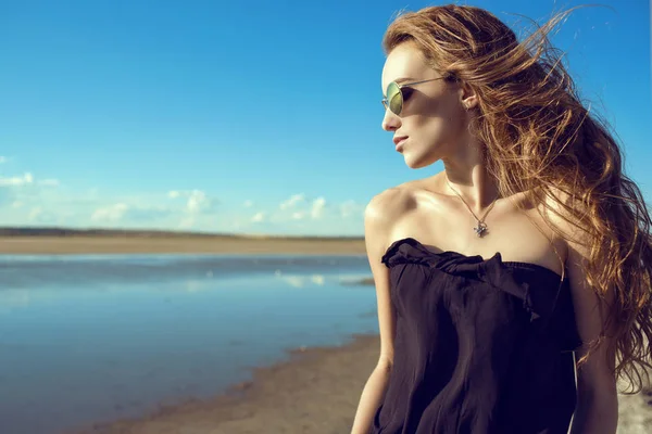 Joven hermosa mujer con estilo con el viento en su pelo con la parte superior del hombro abierto negro y gafas de sol de espejo redondas de moda de pie en la piscina mirando hacia abajo . —  Fotos de Stock