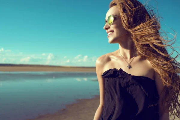 Jovem bela mulher sorridente com o vento em seu cabelo vestindo preto ombro aberto e moda rodada espelhado óculos de sol em pé na piscina olhando para o lado — Fotografia de Stock