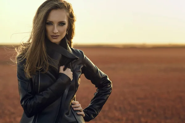 Junges schönes Glam-Model mit dem Wind im langen Haar, das eine schwarze stylische Lederjacke trägt, die bei Sonnenuntergang auf dem verlassenen Feld steht und ihren übergroßen Kragen enger einwickelt — Stockfoto