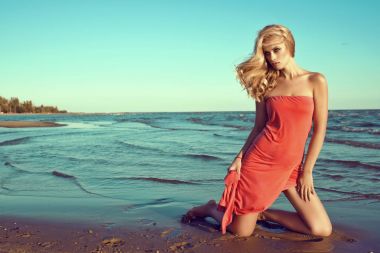 Muhteşem seksi ince sarışın model deniz suyu diz üzerinde duran ve bir kenara elbisesini tren tutan seyir mercan kırmızı straplez elbise. Saçlarını Rüzgar tarafından üflenir