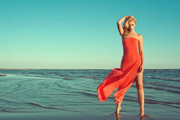 Preciosa modelo rubia delgada sexy en vestido rojo sin tirantes con tren volador de pie en puntillas en el agua de mar, sosteniendo su cabello ondulado soplado por el viento —  Fotos de Stock