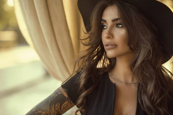 Close up portrait of beautiful tanned glam tattooed model with long wavy hair wearing black dress and wide brimmed hat looking aside — Stock Photo, Image