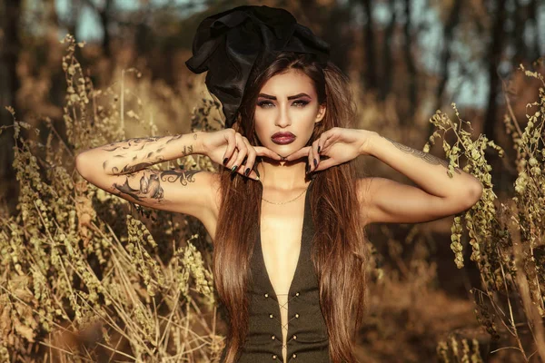Портрет шикарной опасной татуированной ведьмы в сексуальном корсетном платье и винтажной шелковой шляпе, стоящей в лесу и смотрящей прямо с хищным взглядом, ее руки под подбородком. Концепция Хэллоуина — стоковое фото
