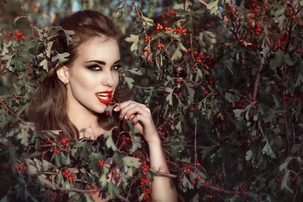 Porträt einer eleganten Frau mit provokantem Make-up und räuberischem Blick, die im Beerenbusch steht und rote Beeren isst — Stockfoto