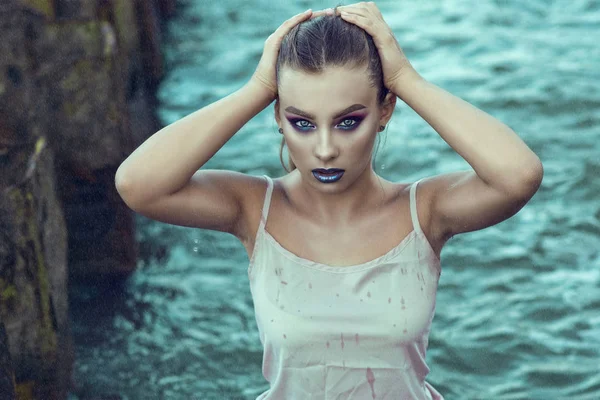 Retrato de una joven hermosa mujer con maquillaje provocativo de pie en el agua de mar bajo la lluvia y tocando su cabello mojado — Foto de Stock