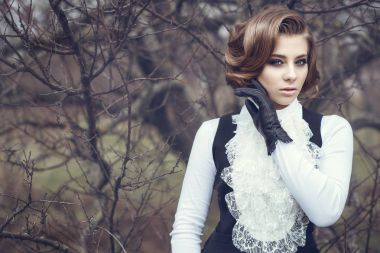 Portrait of gorgeous young woman with elegant Victorian hairstyle and beautiful make up wearing old-fashioned gown with jabot standing at the dry tree holding her hand in leather glove at her cheek clipart