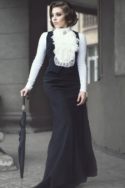 Porträt einer hinreißenden Frau mit eleganter viktorianischer Frisur in einem altmodischen Kleid mit Jabot, die zwischen den Säulen ihres alten Schlosses wandelt und sich auf einen schwarzen Regenschirm stützt — Stockfoto