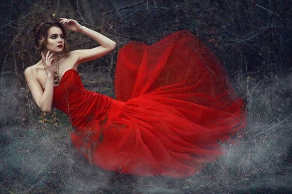 Young beautiful lady with updo hair and provocative make up wearing bright scarlet fish tail dress and vintage necklace sitting in the dry woods covered with fog and looking aside. Fairytale concept