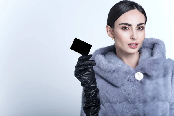 Beautiful smiling dark-haired model with sleek low ponytail wearing blue mink fur coat and long leather gloves holding black blank visit card — Stock Photo, Image