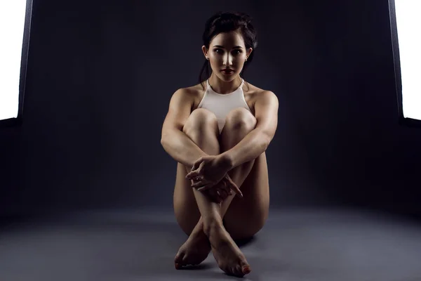 Portrait of young woman athlete in white top sitting on the floor between two studio flash lights embracing her crossed legs