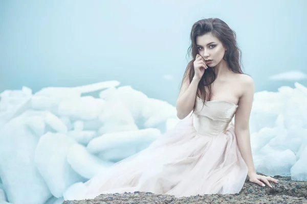 Молодая красивая модель в роскошном корсетном бальном платье без бретелек, сидящая на плитах сломанного льда у туманного моря, рука у губ Лицензионные Стоковые Изображения