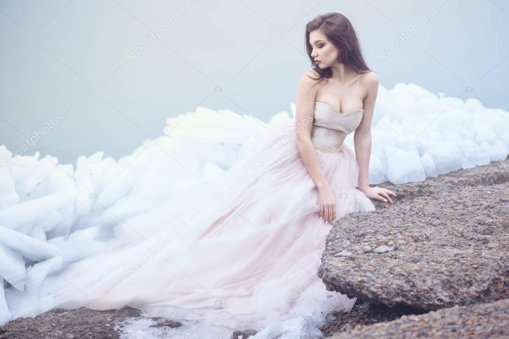 Young beautiful model in luxurious strapless corset ball gown sitting on slabs of broken ice at the misty seaside and looking at the tail of her dress