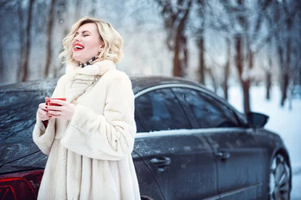 Gyönyörű szőke nő forró kávét iszik a havas téli nap, és nevetve luxus fehér bundát visel csodálatos alkotó — Stock Fotó