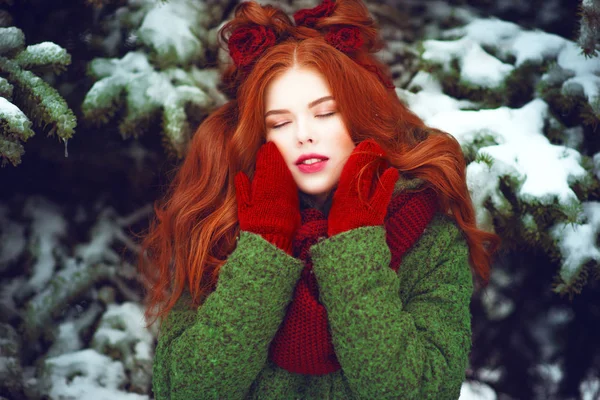 Close up portrait of beautiful red haired girl with creative hairstyle decorated with knitted flowers wearing red knitted mittens and scarf posing with closed eyes in front of snow covered firtrees — Stock Photo, Image