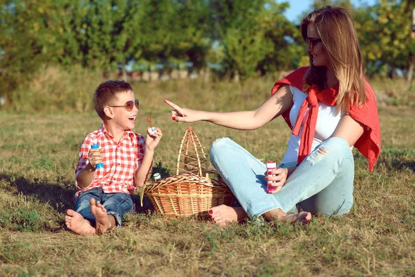 Joven madre elegante divirtiéndose con su pequeño hijo en el picnic al aire libre. Chico está soplando burbujas de jabón y riendo . —  Fotos de Stock