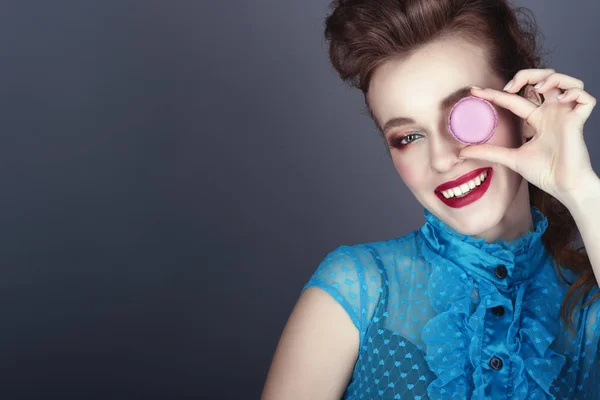 Primer plano retrato de una hermosa modelo con peinado creativo y maquillaje colorido sosteniendo macarrón violeta delante de su ojo y sonriendo — Foto de Stock