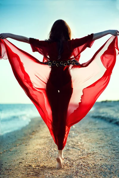 Όμορφη γυναίκα με τέλειο σώμα σε μοντέρνα παραλία κόκκινη Σιφόν καλύψει έως και το περπάτημα στις μύτες των ποδιών κατά μήκος της γραμμής στη θάλασσα προς το ηλιοβασίλεμα κρατώντας τη φούστα της το φόρεμά της που φέρουν στον αέρα — Φωτογραφία Αρχείου