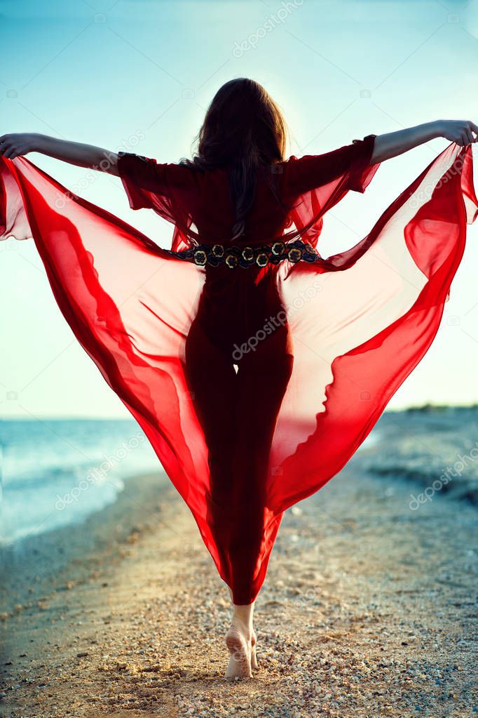 Beautiful woman with perfect body in trendy red chiffon beach cover up walking on tiptoe along the sea line towards the sunset holding the skirt of her gown flying in the wind