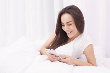 Çok güzel bir genç esmer kadın beyaz yatakta telefonda bir ileti yazar.