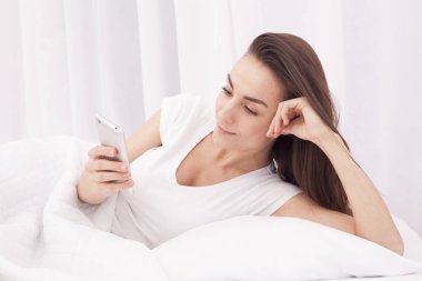 Çok güzel bir genç esmer kadın beyaz yatakta telefonda bir ileti yazar.
