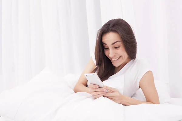 Muito bela jovem morena escreve uma mensagem no telefone na cama branca . — Fotografia de Stock