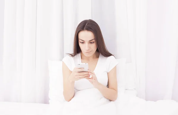 Beautiful young brunette woman writes a message on the phone and listening to music on a white bed.