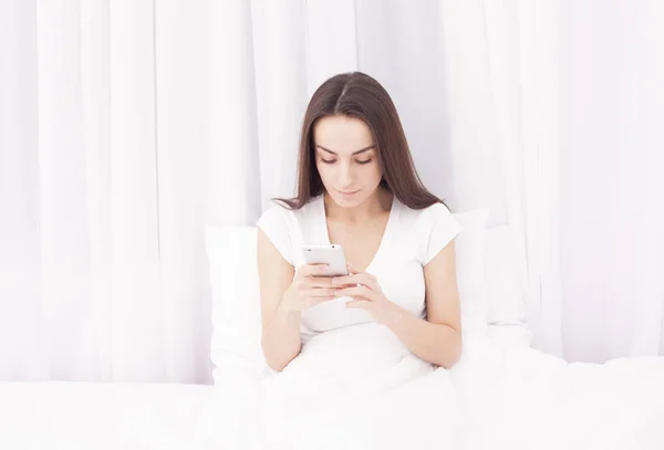 Hermosa joven morena escribe un mensaje en el teléfono y escuchar música en una cama blanca . — Foto de Stock