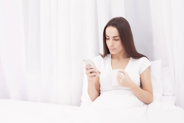 Beautiful young brunette woman writes a message on the phone and listening to music on a white bed.