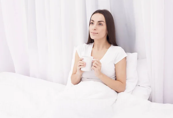 Hermosa joven morena bebiendo café y sonriendo en una almohada blanca . —  Fotos de Stock