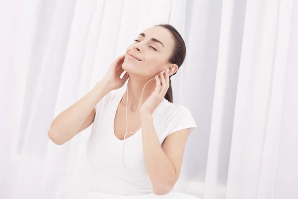 Hermosa chica morena joven divirtiéndose y escuchando música en una cama blanca . — Foto de Stock