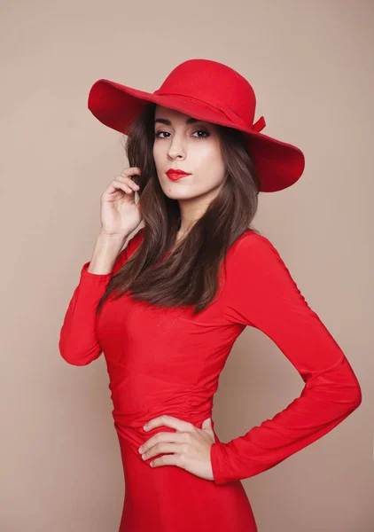 Beauty is in the moment. Unique brunette with red lipstick and elegant red hat on her head in a charming red dress touches a hand to the chin.