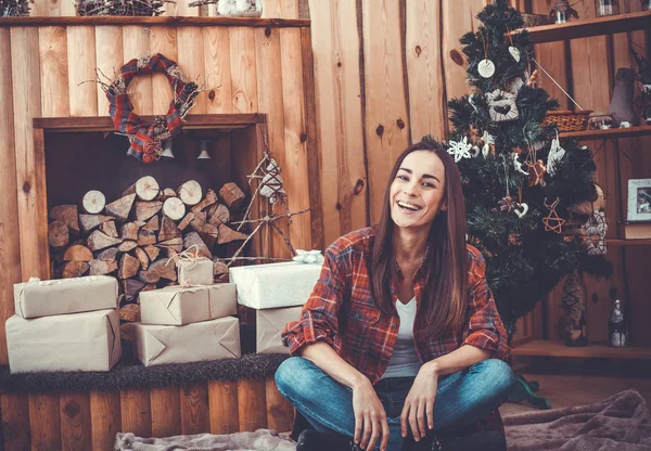 Hermosa joven morena alegre ulybaetsya y sentado en el suelo cerca de un árbol de Navidad y chimenea en una camisa roja casual a cuadros . — Foto de Stock