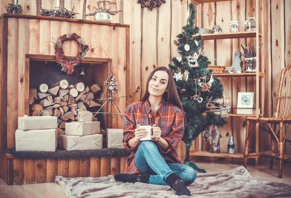 Портрет очень красивой и веселой молодой девушки брюнетки на фоне елки в клетчатой рубашке с красной кофейной кружкой в руках. Новогодняя концепция. Девушка улыбается и отдыхает . — стоковое фото