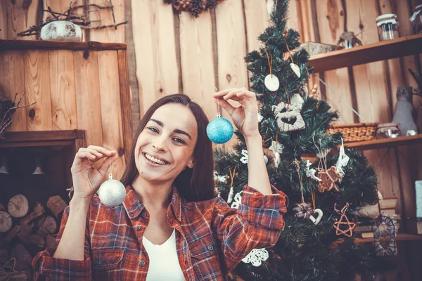 Retrato de una jovencita muy guapa y alegre la morena sobre un fondo del árbol de Navidad en una camisa casual a cuadros con juguetes de Navidad en sus manos. Concepto de Año Nuevo. Sonriendo y descansando . — Foto de Stock