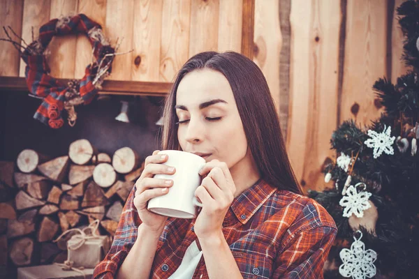 Портрет дуже красивої і веселої молодої дівчини брюнетки на фоні ялинки в плетеній сорочці з червоною чашкою кави в руках. Новорічна концепція. Дівчина посміхається і відпочиває . — стокове фото