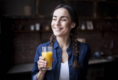 Çok güzel esmer kız evde mutfak kot forması portresi. Gülüyor ve şeffaf bir bardak portakal suyu tutan kız.
