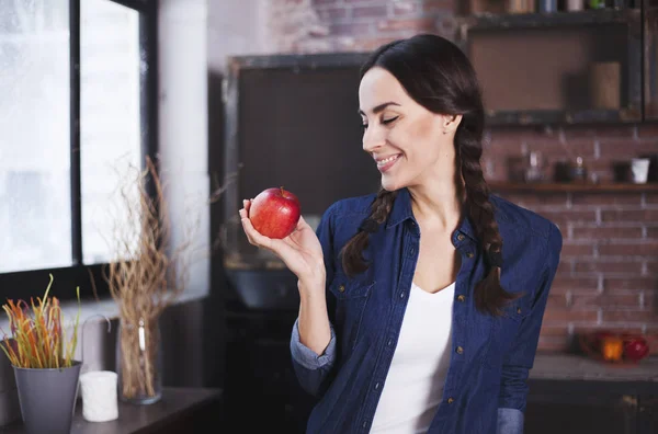 Zeer mooie vrolijke jonge brunette vrouw in denim jasje lacht en toont een rode rijpe appel in haar keuken aan tafel. — Stockfoto