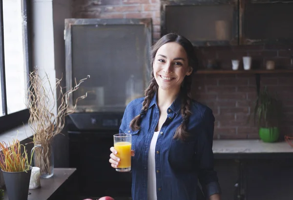 Portrait of very beautiful young brunette girl in jeans shirt on a kitchen at home. Girl laughing and holding orange juice in a transparent glass. — Stock Photo, Image