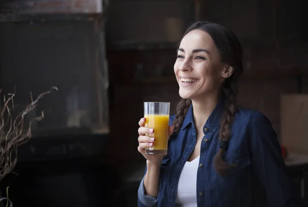 Portret van zeer mooie jonge brunette meisje in jeans overhemd op een keuken thuis. Meisje lachen en jus d'orange te houden in een transparant glas. — Stockfoto