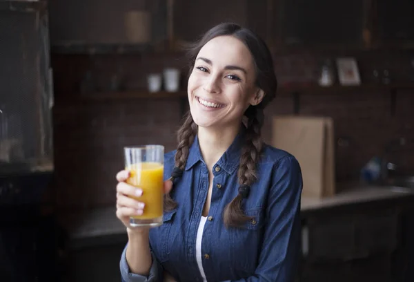 Portret van zeer mooie jonge brunette meisje in jeans overhemd op een keuken thuis. Meisje lachen en jus d'orange te houden in een transparant glas. — Stockfoto
