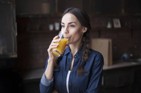 Retrato de jovem morena muito bonita em camisa jeans em uma cozinha em casa. Menina rindo e segurando suco de laranja em um copo transparente . — Fotografia de Stock