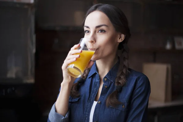 Портрет очень красивой молодой брюнетки в джинсовой рубашке на кухне дома. Девушка смеется и держит апельсиновый сок в прозрачном стакане . — стоковое фото
