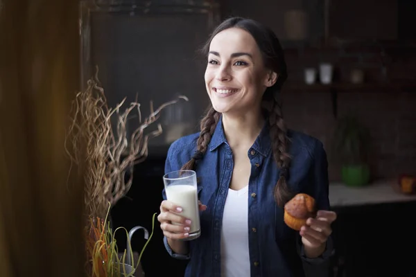 Portret van heel mooi lachende jonge brunette meisje in jeans overhemd op een keuken thuis. Een meisje met een cupcake en melk in een transparant glas. — Stockfoto