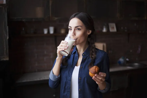 Retrato de jovem morena sorridente muito bonita em camisa jeans em uma cozinha em casa. Uma menina segurando um cupcake e leite em um copo transparente . — Fotografia de Stock