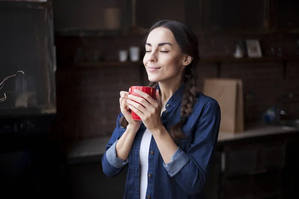 Портрет очень красивой юной брюнетки в джинсовой рубашке с красной чашкой кофе в руках. Девушка улыбается на кухне дома . — стоковое фото